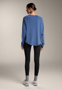 Modra dolga majica s zaobljenim robom, v kombinaciji z črnimi leggingi in sivimi nogavicami vstavljenimi v bele športne čevlje.