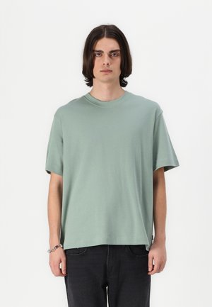 Mintgrünes T-Shirt mit kurzen Ärmeln und Rundhalsausschnitt aus weichem Baumwollstoff, mit einem lässigen Schnitt und seitlichen Nähten. Kombiniert mit dunklen Jeans.