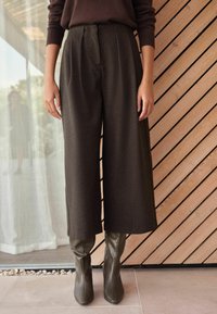 Braune weit geschnittene Hosen mit Falten, aus strukturiertem Stoff. Kombiniert mit olivgrünen, hochhackigen Stiefeln mit quadrater Zehenform. Vor einem Holz-Hintergrund.