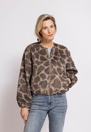 Woman with short blonde hair wearing a brown and gray giraffe-print zip-up jacket and blue jeans, standing against a plain white background.