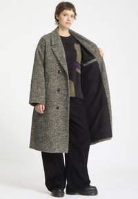 Overdimensioneret sort-hvid flekket frakke med nedskåret revers, to knapper og en lomme indeni. Modellen bærer en mørk mønstret sweater og sorte bukser.