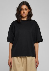 Juoda didelio dydžio marškinėliai iš minkšto medvilnės audinio, su trumpomis rankovėmis ir laisvo silueto, derinami su šviesiai smėlio spalvos kelnėmis.