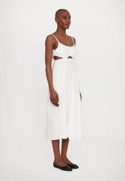 Vestido maxi blanco con tirantes finos, que presenta detalles recortados en la cintura, tela fluida y una textura suave; combinado con bailarinas negras.
