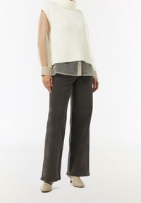 Vrouw in een beige mouwloze gebreide vest over een doorschijnende blouse met lange mouwen, donkere broek met brede pijpen en beige haklaarzen, staand.