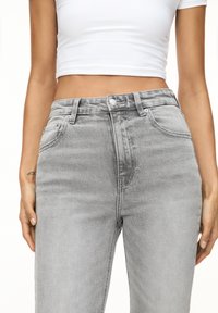 Femme portant un jean taille haute gris clair et un haut court blanc, les mains détendues le long du corps, devant un fond uni.