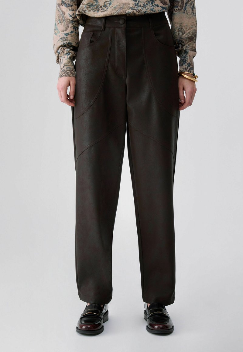 Personne portant un pantalon marron foncé taille haute avec poches latérales, associé à une chemise à manches longues à motifs et des mocassins noirs brillants.
