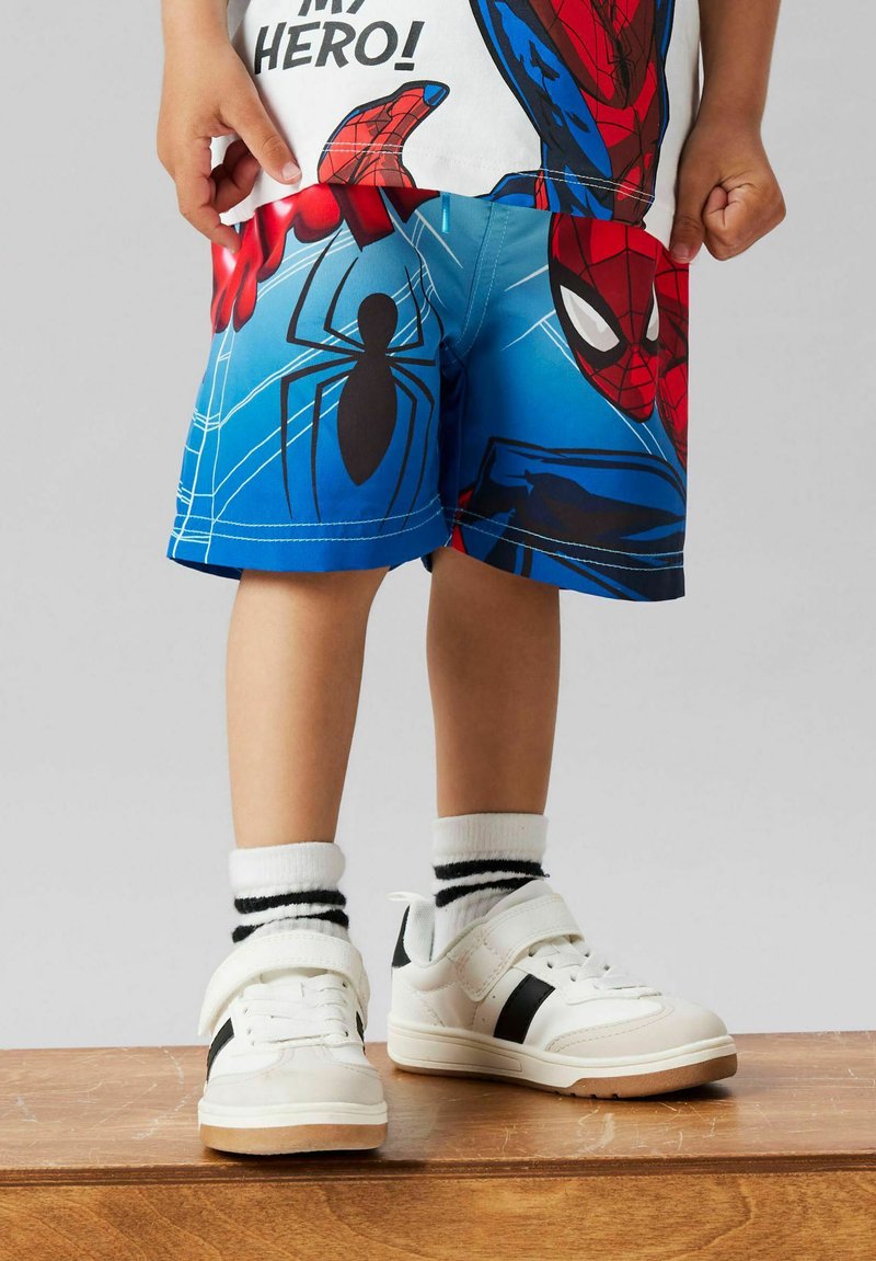 Enfant portant un short bleu Spider-Man, des chaussettes blanches à rayures noires et des baskets blanches, debout sur une surface en bois.