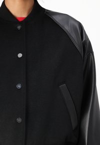 Čierna varsitná bunda s vlneným telom a koženými rukávmi, okrúhlym golierom, predným zapínaním na gombíky a bočnou kapsou s elegantným dizajnom.