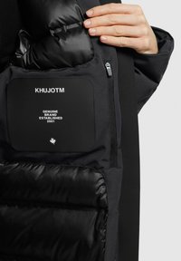 Black insulated jacket featuring a smooth outer layer, quilted lining, and a patch with "KHUJOTM" text and logo on the inner pocket.