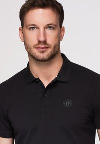 Jeune homme aux cheveux bruns courts et barbe taillée portant un polo noir avec un petit logo brodé sur la poitrine sur un fond uni.