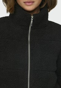 Zwarte fleece jas met hoge kraag, voorzien van een zilveren rits aan de voorkant. Gelaagd stof met een zachte, pluche uitstraling.
