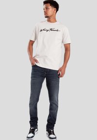 Camiseta gris claro con "Wrong Friends" impreso en cursiva negra. Combinada con jeans de denim oscuro y zapatillas en blanco y negro.