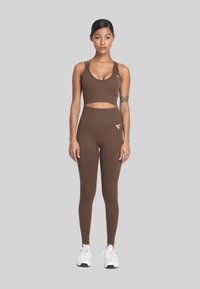 Kobieta stoi prosto, ubrana w brązowy stanik sportowy i legginsy oraz białe sneakersy na tle jednolitego szarego tła.