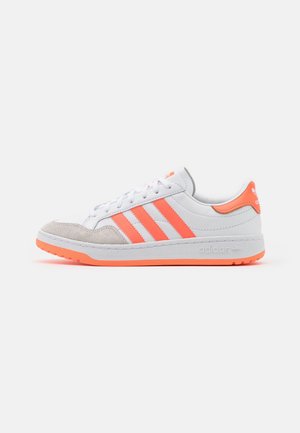 Baskets basses blanches avec des rayures orange, semelle orange, bout en daim et languette de talon orange portant le logo Adidas, vues de profil.