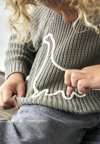 Pilkas megztinis su kreminės spalvos dinozaurų kontūro dizainu, derinamas su pilkomis kelnėmis. Medžiaga turi tekstūruotą, grublėtą išvaizdą.