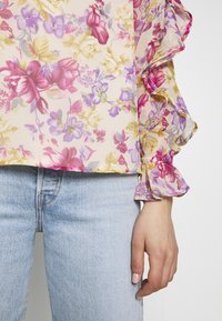 Blouse fleurie transparente avec poignets à volants dans des tons roses, violets et jaunes, associée à un jean en denim bleu clair.