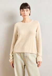 Beige kötött pulóver, texturált, puha felülettel, kerek nyakkivágással és hosszú ujjakkal. Világoskék, kényelmes szabású farmernadrággal párosítva.