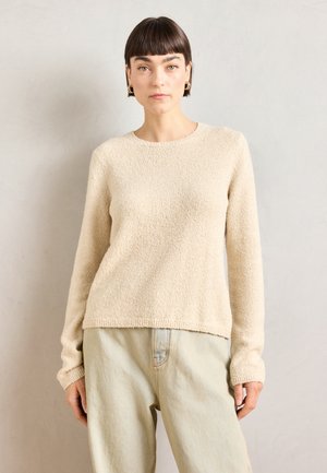 Pull en tricot beige avec une surface texturée et douce, encolure ronde et manches longues. Assorti à un jean bleu clair à coupe décontractée.