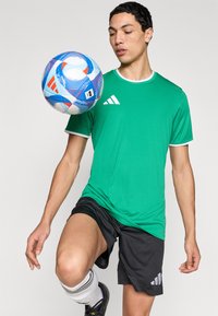 Πράσινο αθλητικό πουκάμισο με λευκή επένδυση, μαύρο σορτς και πολύχρωμη ποδοσφαιρική μπάλα με σχέδιο που ισορροπείται σε πόδι.