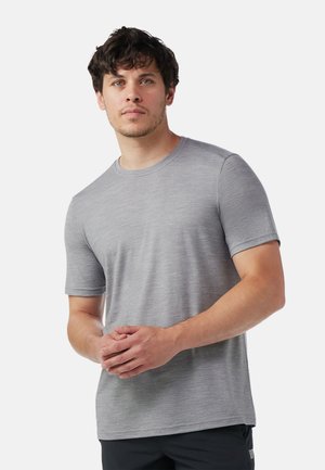 Mand med kort krøllet hår iført en simpel lysegrå kortærmet t-shirt og sorte bukser, stående med hænderne foldet.