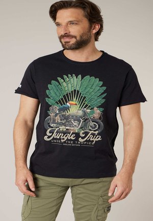 Homme portant un t-shirt noir avec un motif de moto, feuilles tropicales, toucan, singe, et texte "Jungle Trip under the Tropics" en lettres beiges.