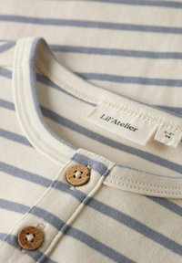 Pasiasty top z bawełny w jasnobeżowe i niebieskie poziome paski, okrągłym dekoltem oraz dwoma drewnianymi guzikami. Metka głosi "Lil'Atelier."