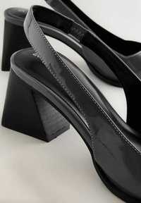 Escarpins slingback en cuir verni noir avec talons carrés sur un fond clair, montrant les détails en gros plan des sangles et des coutures.
