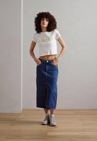 T-shirt blanc raccourci avec un design graphique vert, associé à une jupe longue en denim bleu avec une fente avant et des baskets montantes grises.