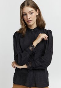 Blusa nera in tessuto testurizzato, con dettagli in pizzo sul colletto e sui polsini, chiusura con bottoni al collo e maniche ampie.