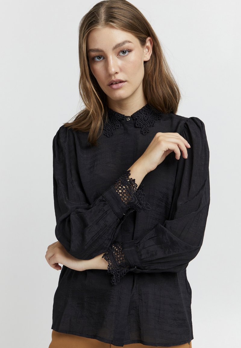 Blusa nera in tessuto testurizzato, con dettagli in pizzo sul colletto e sui polsini, chiusura con bottoni al collo e maniche ampie.