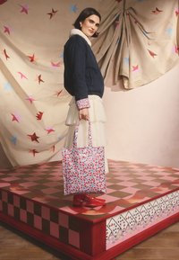 Baltas gėlių raštuotas rankinės tipo krepšys su raudonomis gėlėmis, laikomas asmens, dėvinčio tamsiai mėlyną švarką ir sluoksniuotą kreminės spalvos sijoną, stovinčio ant rožinės languotos platformos.