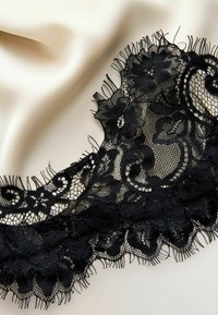 Tissu en dentelle florale noire avec des motifs complexes et des bords à franges sur un fond en satin crème lisse.