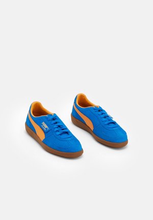 Paire de baskets Puma Palermo bleues avec une bande orange et une doublure intérieure, semelles marron et lacets, présentée sur fond blanc.