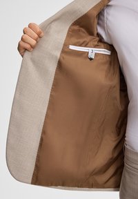 Heller Blazer mit einer strukturierten Außenseite und glattem braunem Innenfutter. Verfügt über eine Innentasche mit Knopfverschluss.