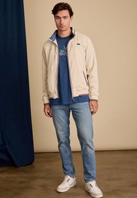 Veste beige clair à col montant, doublure bleue et poignets à rayures contrastées ; portée avec un t-shirt graphique marine et un jean bleu clair.
