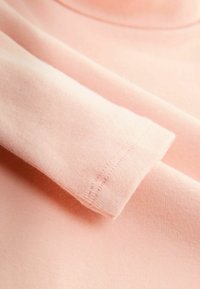 Lyserød bomuldsstof med en glat overflade, der har en rullet ærme med en ren søm. Stoffet virker blødt og let strækbart.