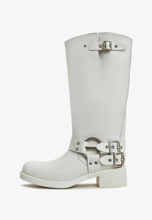 Botte blanche jusqu'au genou en matériau lisse, ornée de boucles métalliques brillantes et d'un accent circulaire, avec un talon rectangulaire et une semelle texturée.