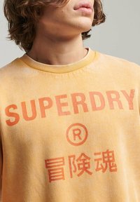 Žlutá mikina s vybledlou texturou, na přední straně potisk s logem značky "SUPERDRY" a čínskými znaky v oranžové barvě.