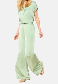 Jumpsuit di seta verde chiaro con maniche corte, vita aderente e design a gamba larga. Presenta una lucentezza sottile e una vestibilità rilassata, abbinato a sandali beige.