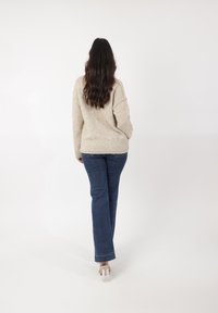 Beiger Strickpullover mit lockerer Passform, kombiniert mit dunkelblauen Schlagjeans und transparenten High Heels, von hinten betrachtet.