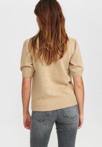 Beżowy sweter z dzianiny z krótkimi rękawami, ze ściągaczem na dole i przy rękawach, o gładkiej fakturze. W połączeniu z szarymi dopasowanymi dżinsami.