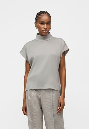 Kvinne med flettefrisyre iført store hoop-øredobber, en lys grå kortermet polotrøye og grå rutete bukser med vide ben, stående foran en ensfarget bakgrunn.