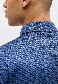 Κοντινό πλάνο του λαιμού και του ώμου ενός άνδρα που φοράει ένα μπλε πουκάμισο με γιακά και σχέδιο, με ένα μικρό μαύρο λογότυπο στο πίσω μέρος.