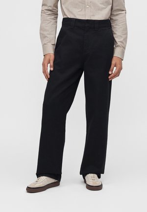Homme portant un pantalon noir droit, une chemise beige à manches longues et des baskets beiges, debout sur un fond blanc uni.