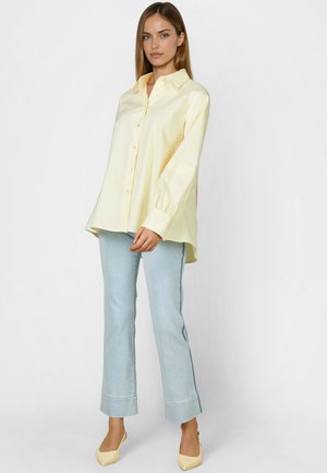 Woman wearing a loose pale yellow button-up shirt, light blue wide-leg jeans, and pale yellow pointed-toe heels, standing against a plain background.
