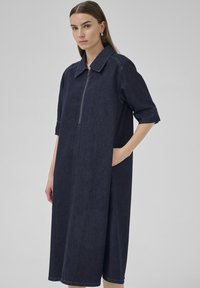 Temna denim obleka s kratkimi rokavi, sprednjim zadrgo in stranskimi žepi, s strukturiranim ovratnikom in dolžino do kolen.