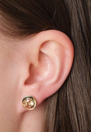 Nahaufnahme eines linken Ohrs mit braunem Haar, das einen runden goldenen Ohrstecker mit einem hellorangen Edelstein trägt.