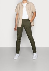 Beige T-shirt met korte mouwen over een wit T-shirt, gecombineerd met olijfgroene slim-fit broek en witte sneakers; soepele texturen en een eenvoudig design.
