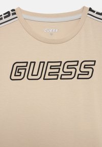 Beige t-shirt met een ronde hals, voorzien van een groot zwart "GUESS" logo en zwart-witte gestreepte accenten op de schouders. Zachte textuur.