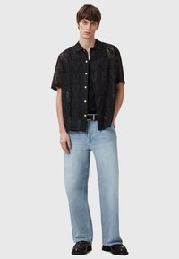 Camisa negra de manga corta con un intrincado patrón de cortes, combinada con jeans de pierna ancha en azul claro y zapatos de vestir negros, presentada sobre un fondo neutro.